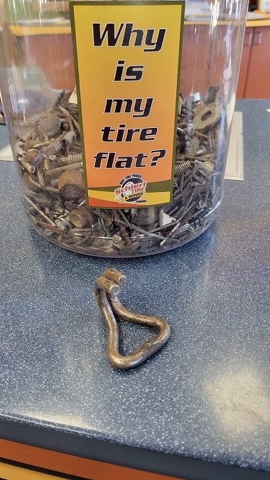 Container with screws, nails, and miscellaneous objects that have caused flat tires. In front of the container is a broken, metal mystery object that a technician pulled from a punctured tire